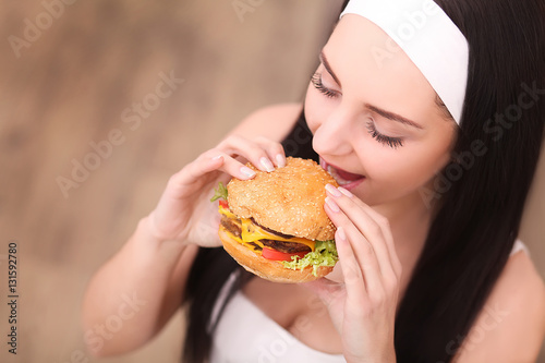 Unhealthy eating. Junk food concept. Portrait of fashionable young woman holding burger and posing over wood background. Close up. Copy-space. Perfect hair, skin, make-up and manicure. Studio shot