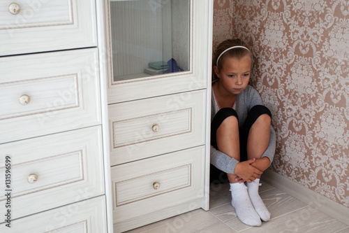 Sad little girl sitting in the corner of a room behind the cupboard