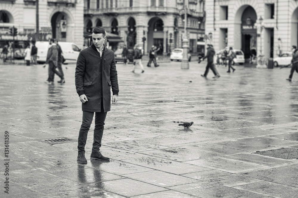 Full length of young man in coat walking under rain along square in European city. Turin center, Italy