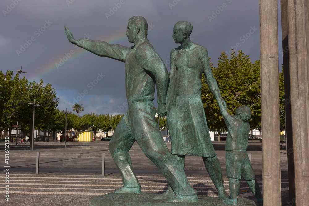 A statue commemorating migrants in Ponta Delgada, Azores, Portugal. The monument and rainbow across low stormy skies on Sao Miguel Island in Azores archipelago.