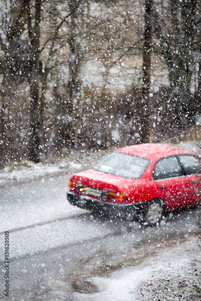 A red car is driving in a snow blizzard. The car is out of focus. Dangerous weather to drive. Heavy snowfall on a winter day.