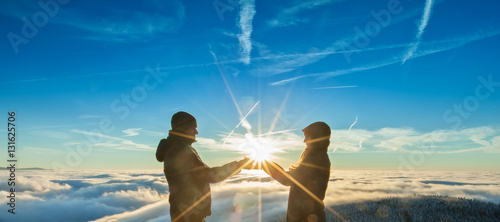 Φωτογραφία The Silhouette of man and woman with sun in hands standing on the top of mountai
