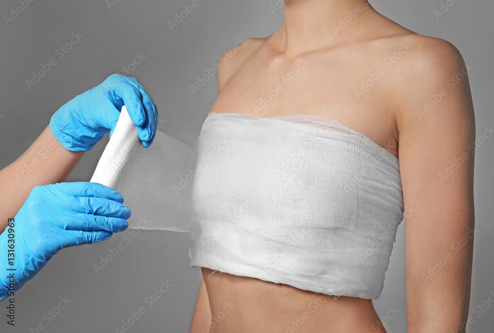 Doctor wrapping bandage around young woman's chest on grey background,  closeup Stock Photo