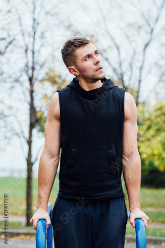 portrait of focused muscular young man in black workout clothes doing dips on parallel bars.