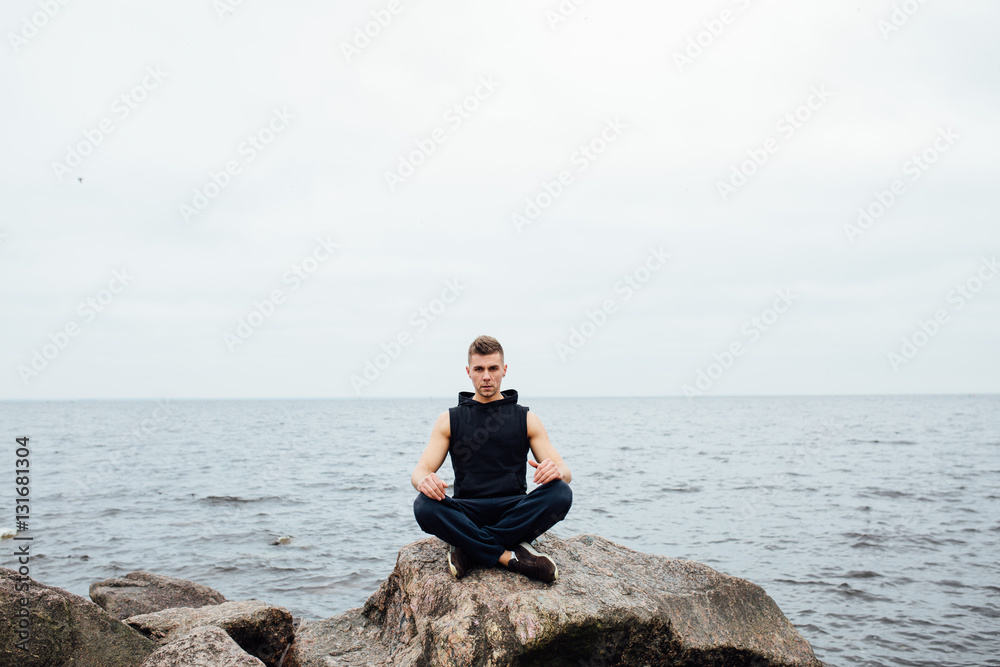 Strong fitness yoga man in lotus pose on the rock beach near the ocean. Harmonic concept.