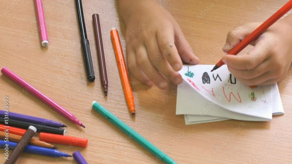 Child sitting at the desk draws the picture on the paper sheet using the colored felt pens. Close-up Child sitting at the desk draws the picture on the paper sheet using the colored felt pens. Close-up