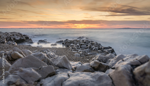 The sun sets over the beach in Petrčane, Croatia, with steely colored rocks in the foreground and the waves of the Adriatic Sea misty in the evening light with a long exposure effect.