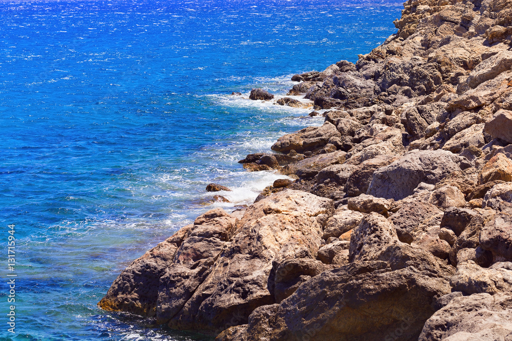 Stones along the seashore. Blue seawater. Small waves beating against the shore. Unapproachable deserted stony shore in the region Lenta on the island of Crete