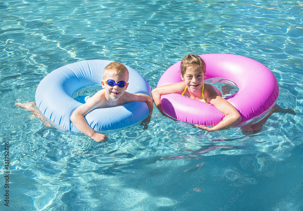 Two cute kids playing on inflatable tubes in a swimming pool on a sunny day.  Bright colorful fun summer photo with copy space Photos | Adobe Stock