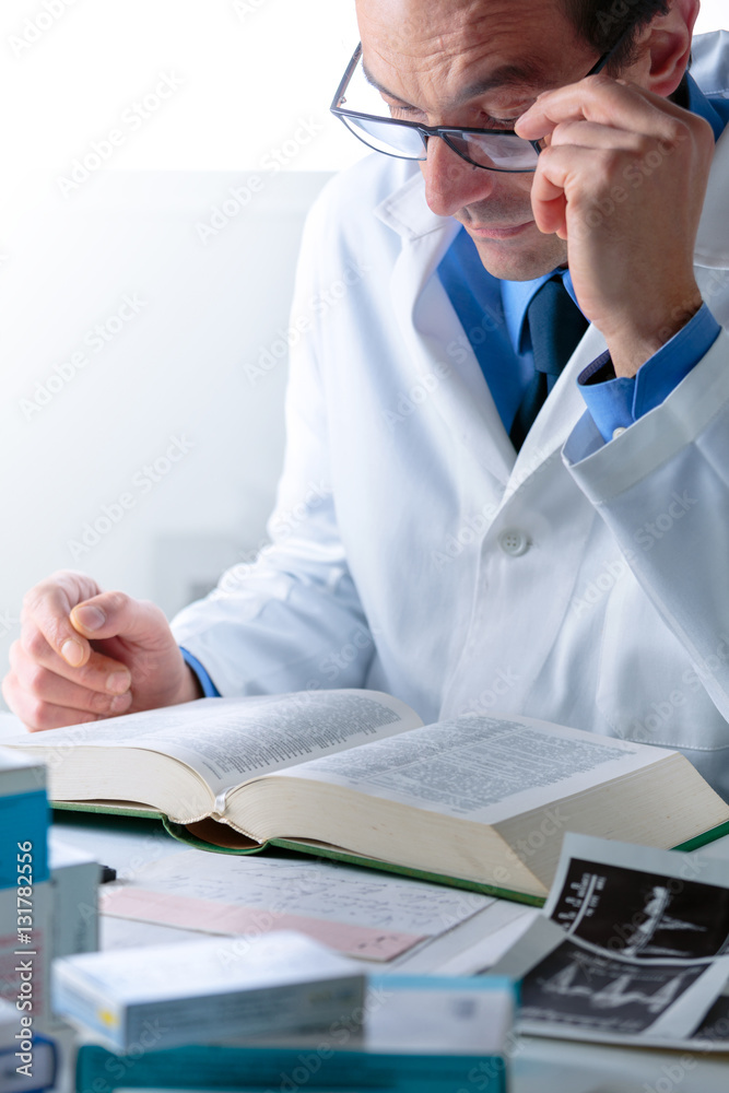 Senior Male Doctor looking for Information in a Medical Book at Senior Male Doctor looking for Information in a Medical Book at