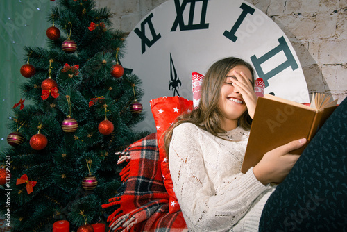 Beautiful girl reading a book lying on the couch, covered with plaid. Winter holidays, New Year, a large clock. Interior room 
