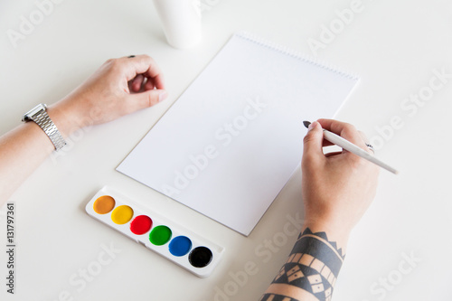 Man's Hand with a tattoo hold the brush in front of blank paper and paints on white background. Branding Mock-Up.