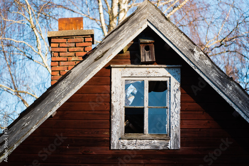 Broken window on an old red wooden house. Birdhouse or nesting box above window on gable. Smokestack in background. © imfotograf