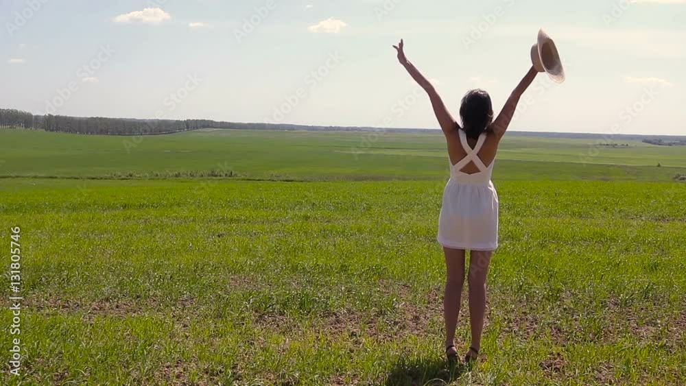young girl with long dark hair and standing throws hands with straw hat on a green field with green grass backs to the camera in a short white summer dress and his hair. young girl with long dark hair and standing throws hands with straw hat on a green field with green grass backs to the camera in a short white summer dress and his hair.