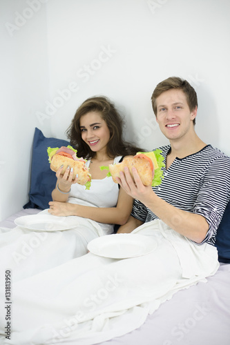 Happy couple lying in bed in morning and holding hamburgers for eating. Happy man and woman prefer having morning breakfast in bed.