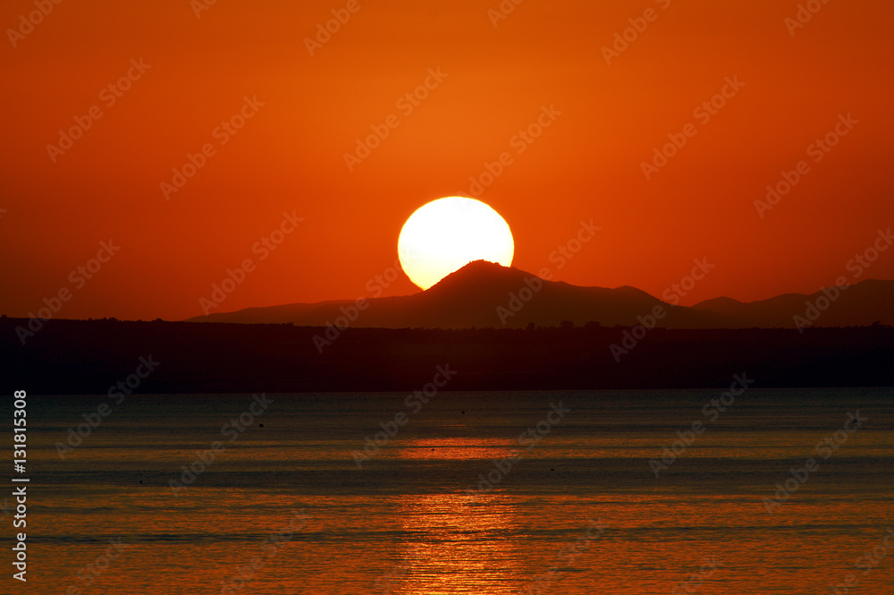 The sun against the background of the red sky sets behind the mountain and is reflected in the sea.