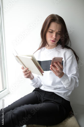 Irritated woman sitting on windowsill and looking down to smartphone