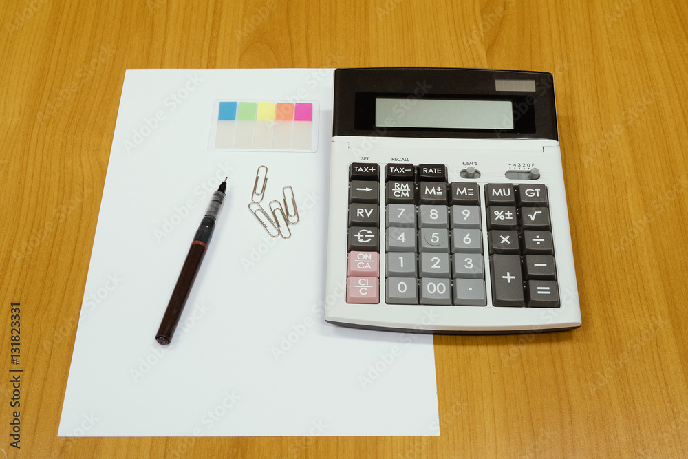 Office equipment, a pen, a calculator, a page marker and clips on a plain white paper with a wood desk as background Office equipment, a pen, a calculator, a page marker and clips on a plain white paper with a wood desk as background