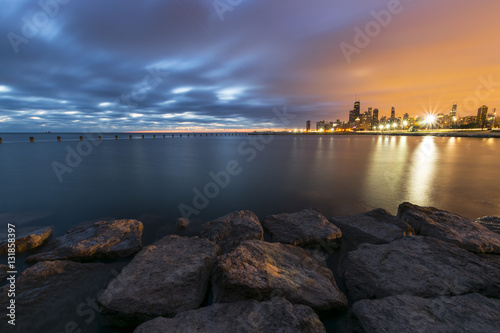 A shot of an empty lake taken from a pier during the sunrise with Chicago's Skyline visible in the background. Beautiful rocks are present in the foreground. .