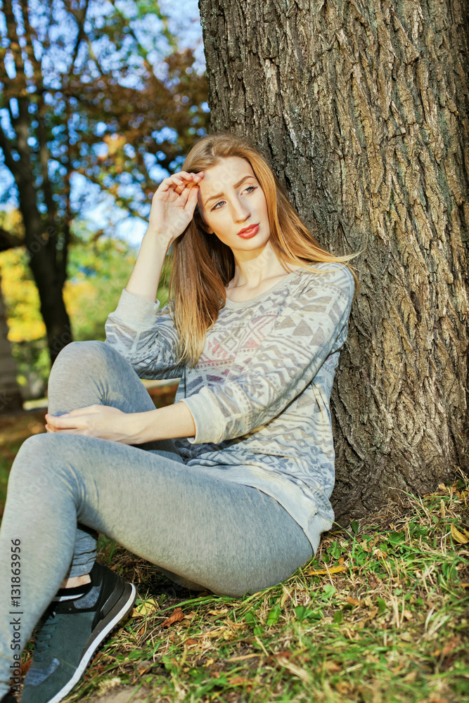 Young beautiful girl with blue eyes and blonde hair sitting near a tree Young beautiful girl with blue eyes and blonde hair sitting near a tree