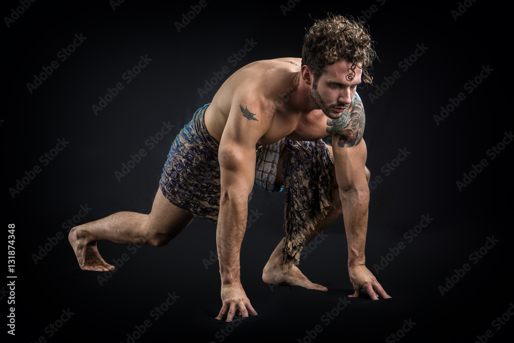 Handsome shirtless muscular man in briefs, covering with long elegant scarf, kneeling, isolated on black background in studio shot