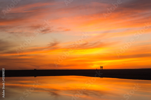 orange and yellow clouds at sunset on a beach with people run and sky reflected in water