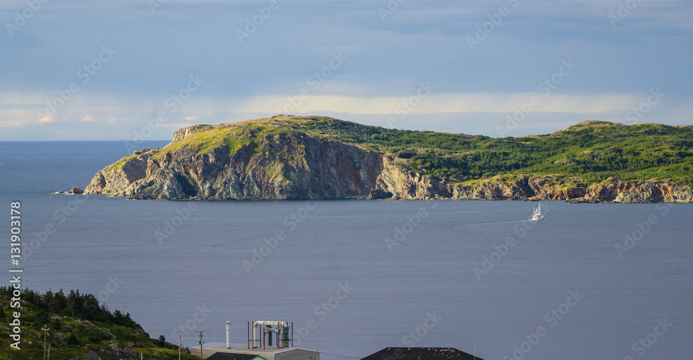 Φωτογραφία Late afternoon sun shines on an outcropping of high rugged terrain in calm sea water as a sailboat under motor power, goes along in the bay at Twillingate, Newfoundland, Canada