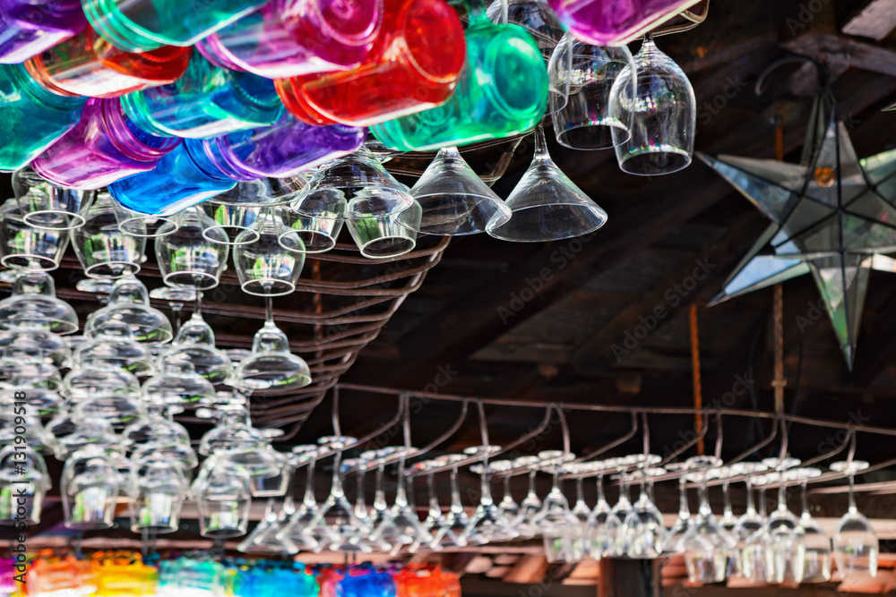 Restaurant interior background with narrow focus. Empty transparent wineglasses, rows of colorful beverage glasses hanging on top rack above bar counter. Vintage style wood ceiling.