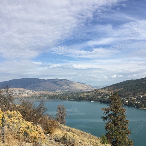 Fototapeta Naklejka Na Ścianę i Meble -  Scenic lake landscape with blue water, mountains,blue sky and white cloud patterns.Tall green tree and bushes isolated on hillside by lake in autumn. Yellow dry grass on hill and sunshine on mountain.