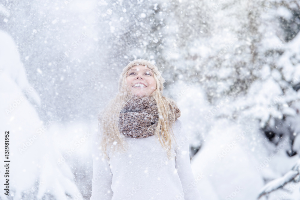 Attractive smiling young blonde girl walking in winter forest. Pretty woman in wintertime outdoor. Wearing winter clothes. Knitted sweater, scarf, hat and mittens.