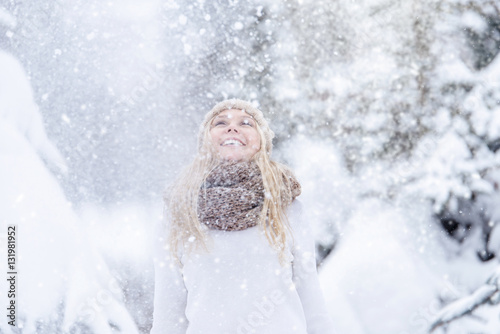 Attractive smiling young blonde girl walking in winter forest. Pretty woman in wintertime outdoor. Wearing winter clothes. Knitted sweater, scarf, hat and mittens.
