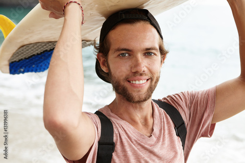 People, hobby and leisure concept. Happy young bearded surfer holding surfboard over his head looking and smiling cheerfully at camera, can't wait to go surfing in perfect weather conditions