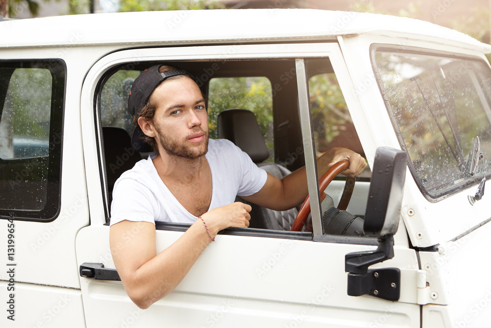 Close up shot of fashionable handsome young bearded model posing inside white jeep on driver's seat holding hand on steering wheel and looking at camera with confident expression on his face