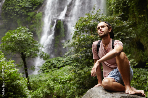 People  nature and adventure. Young hipster with backpack sitting on big rock by waterfall on background  having joyful look. Stylish barefooted hiker relaxing on stone during journey in jungle