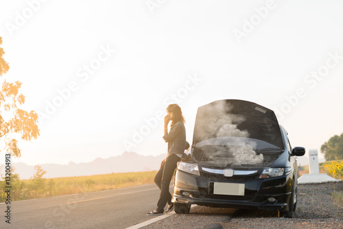 Broken car. Lady standing on the road by the broken car in the middle of nowhere. Making telephone call to get help with the broken car. smoke coming out the engine. Help needed. Car service. 