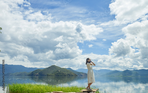 Single young Chinese woman in white long skirt stands on wasted platform on bank of Lugu lake, listening to music, back to camera, at Daluoshui village, Lijiang, China photo