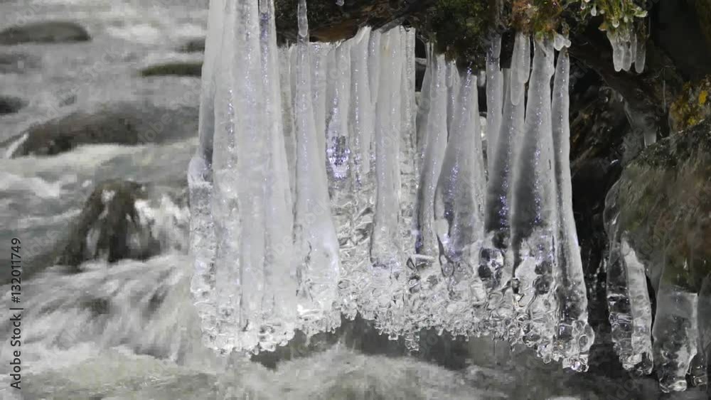 Long icicles hang above dark cold water of mountain river. Icicles glitter above milky water of stream. Branches of fallen trunks are covered by small flakes of powder snow. Long icicles hang above dark cold water of mountain river. Icicles glitter above milky water of stream. Branches of fallen trunks are covered by small flakes of powder snow.