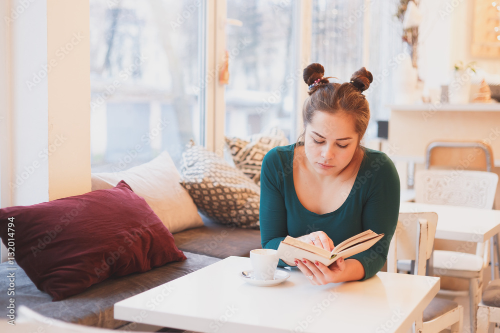 Portrait of attractive young girl enjoying a good book and cup coffee sitting in comfortable interior.