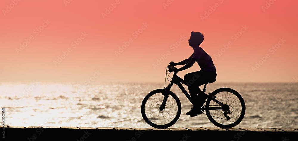 Obraz premium Silhouette of young boy cyclist on sunset, along seashore 