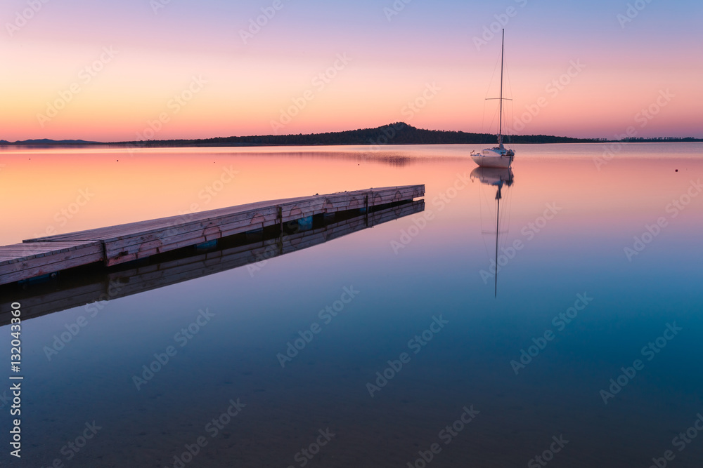 Sunset over lake with yacht, moored by wooden pier.