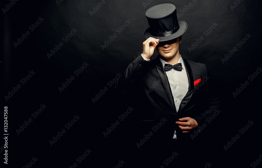 A magician in a black suit holding an empty top hat and magic wand isolated on black background - obrazy, fototapety, plakaty 