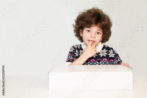 Thoughtful cute young boy in a sweater with an ornament biting her finger, stares into the camera. Holding in hands a white box. Gray background.