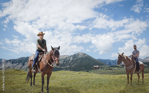 horizontal image of a father and teenage son out horse back riding as they sit on their horse and pose for camera with mountains and blue sky over head in the summer time.