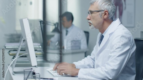Wallpaper Mural Senior Medical Practitioner Working in His Office at His Desktop Computer. Also His Assistant Works in the Background.  Shot on RED Cinema Camera in 4K (UHD).  Torontodigital.ca