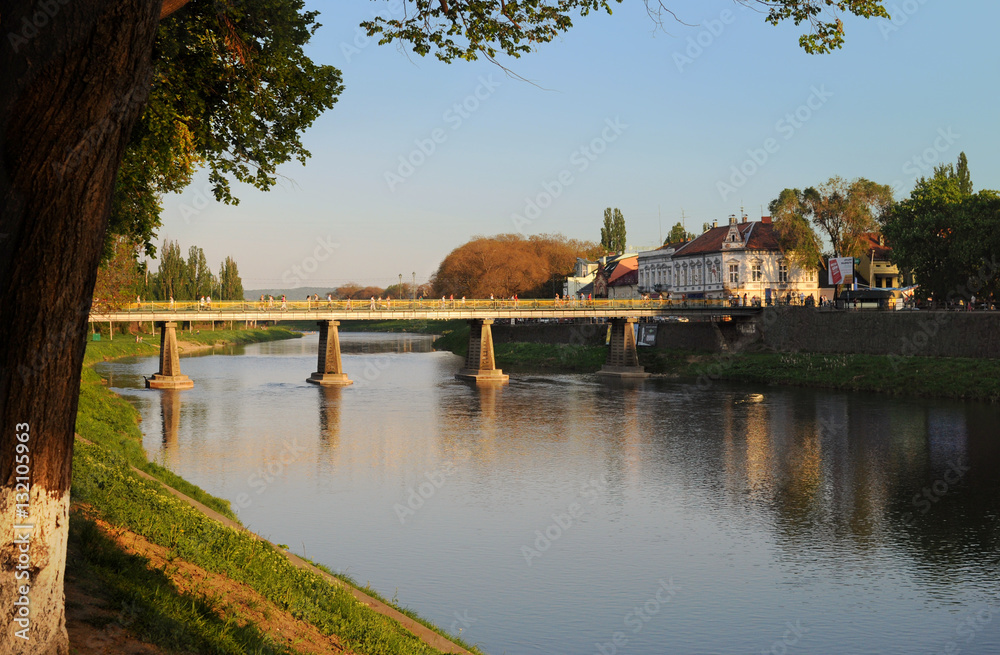 View on the main bridge across the river Uzh in Uzhhorod in warm sunset light. High tree grows on the left View on the main bridge across the river Uzh in Uzhhorod in warm sunset light. High tree grows on the left