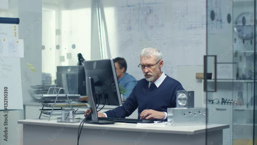 Busy Engineer Bureau. Chief Engineer Working at His Desk, Some Technical Components Lying on His Desk. His Assistant Works at His Desk in the Background. Shot on RED Cinema Camera in 4K (UHD). Busy Engineer Bureau. Chief Engineer Working at His Desk, Some Technical Components Lying on His Desk. His Assistant Works at His Desk in the Background. Shot on RED Cinema Camera in 4K (UHD).
