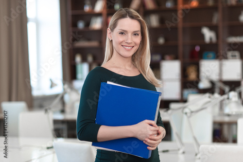 Woman standing in office and holding a folder in his hands. Looking at the frame and smiles.