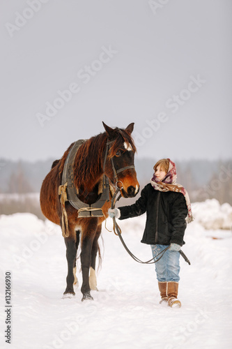 Little young girl in winter on the snow in a fur coat and headscarf leads the horse by the bridle and looking at it