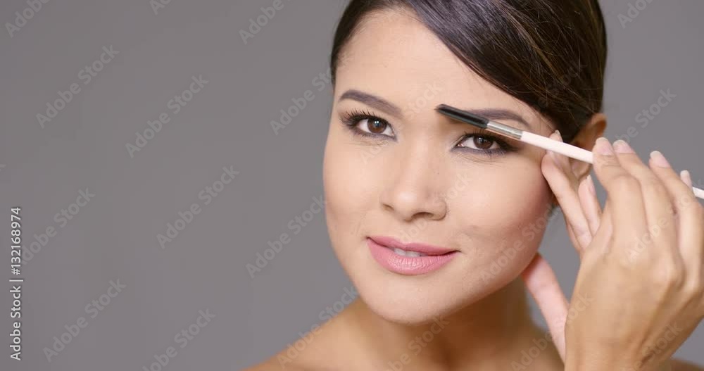 Young woman darkening her eyebrows applying makeup with a cosmetics brush in a close up cropped beauty facial portrait on grey with copy space Young woman darkening her eyebrows applying makeup with a cosmetics brush in a close up cropped beauty facial portrait on grey with copy space