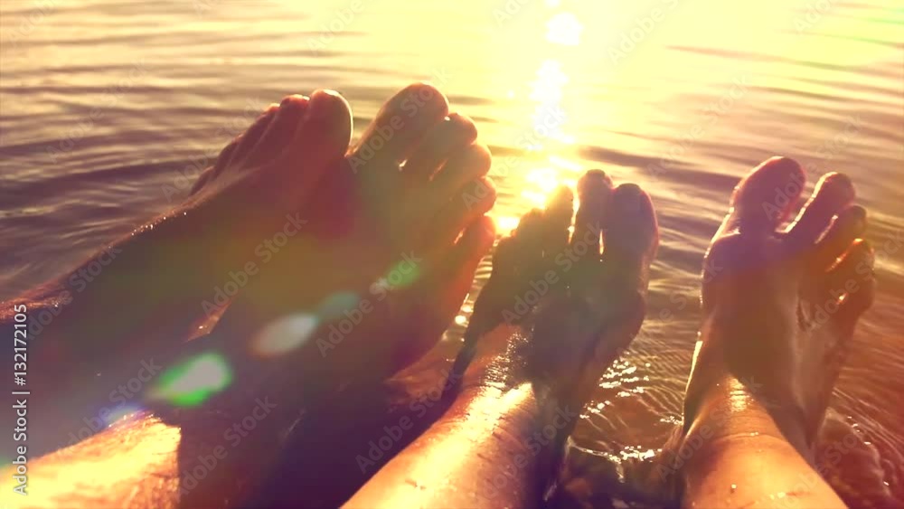 Summer holidays, Vacation concept. Bare feet of young man and woman in the water. Relaxing couple on the beach. Over summer sunset. Sun flare. Slow motion 1080p video footage